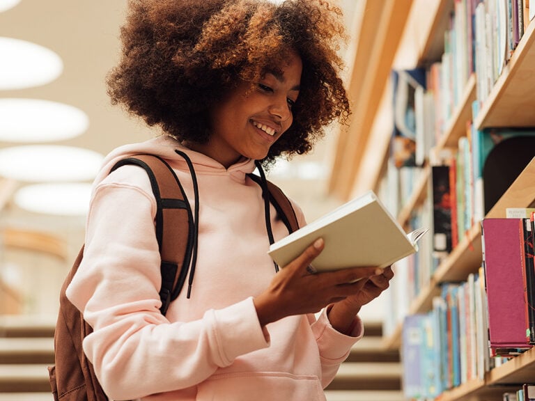 Smiling girl holding a book in library while standing at bookshelf