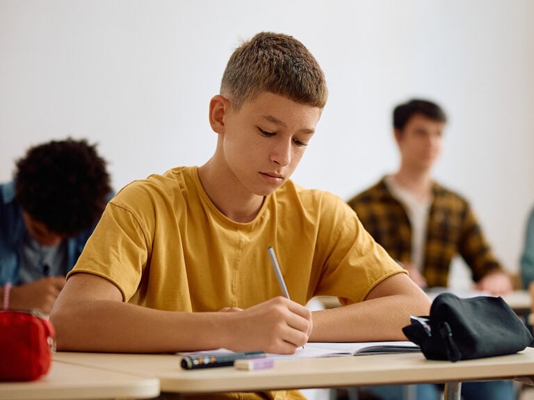 Male high school student writing in notebook in classroom.